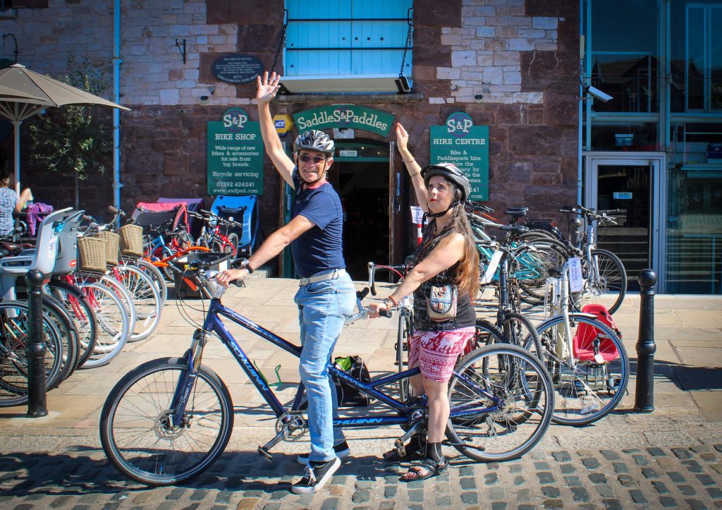 A photo of Dawn and her tandum bike riding partner, sitting on the tandem bike outside Saddles & Paddles Bike Hire Centre, about to set off on their sponsored bike ride. 