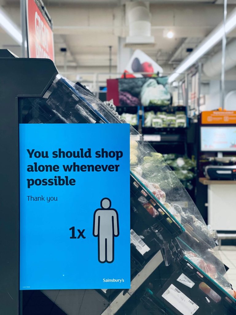 The photo shows a blue sign in a Sainsbury's store that says "You should shop alone whenever possible - Thank you" with an icon of a single person and Sainsbury's on the bottom of the sign. The sign is next to boxes containing apples and there are other fruit and veg in the background.